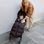 A woman in a brown jacket and beige pants pushes a stroller while a baby sits inside the Finnson alma universal footmuff in chocolate leopard. The scene is set outdoors against a light gray tiled wall and stone floor.