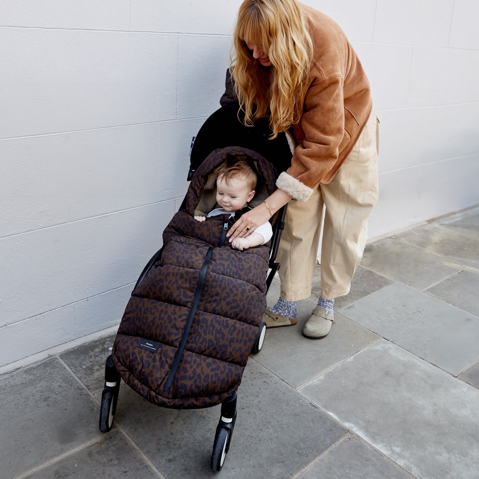 A woman in a brown jacket and beige pants pushes a stroller while a baby sits inside the Finnson alma universal footmuff in chocolate leopard. The scene is set outdoors against a light gray tiled wall and stone floor.