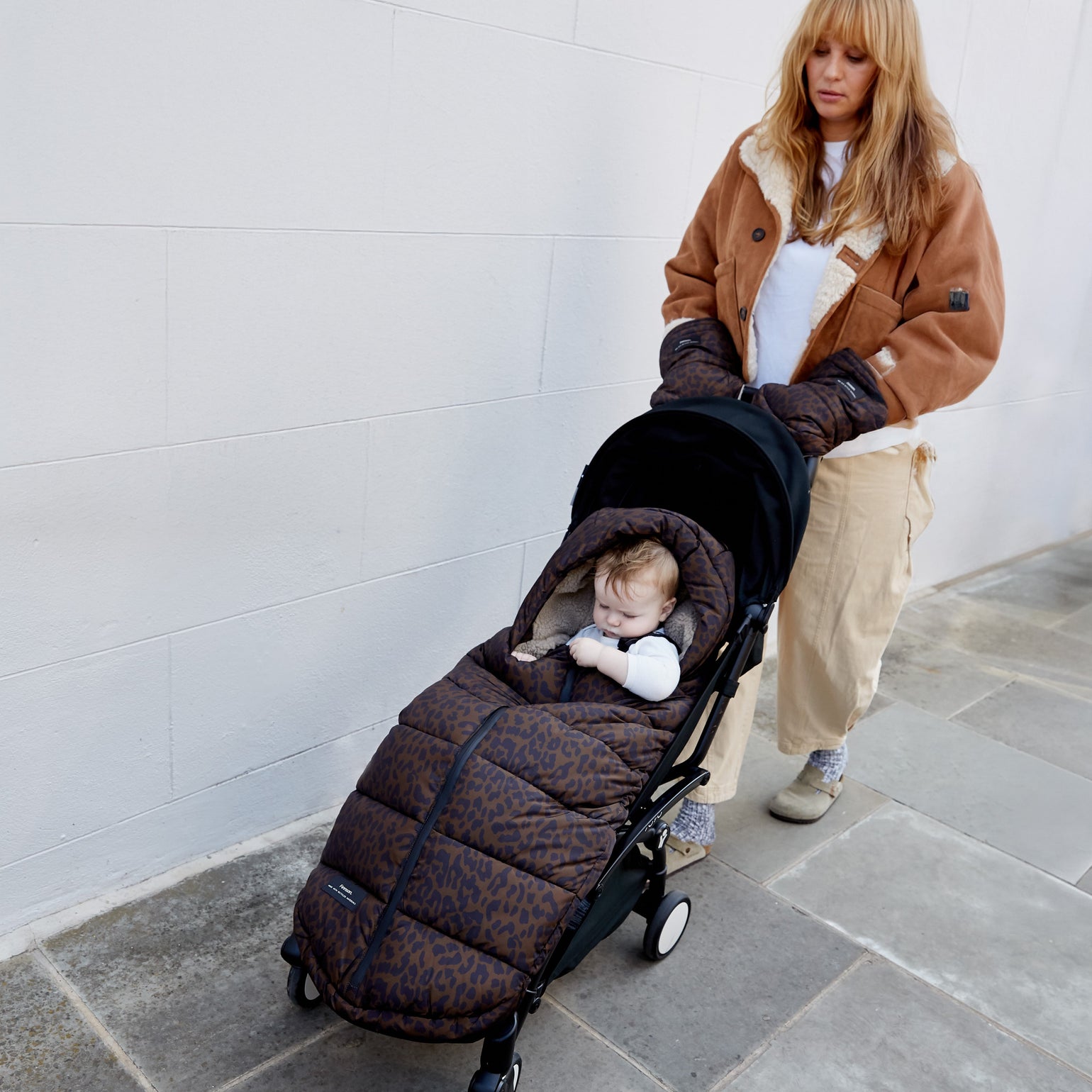 A woman pushes a stroller on a stone paved walkway against a white wall. The stroller is covered with the alma universal footmuff in chocolate leopard, and a baby is sleeping inside.