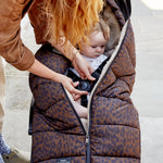 The alma foot muff in chocolate leopard  with a beige fleece lining, shown on a stroller with a baby inside and an adult adjusting the zipper.