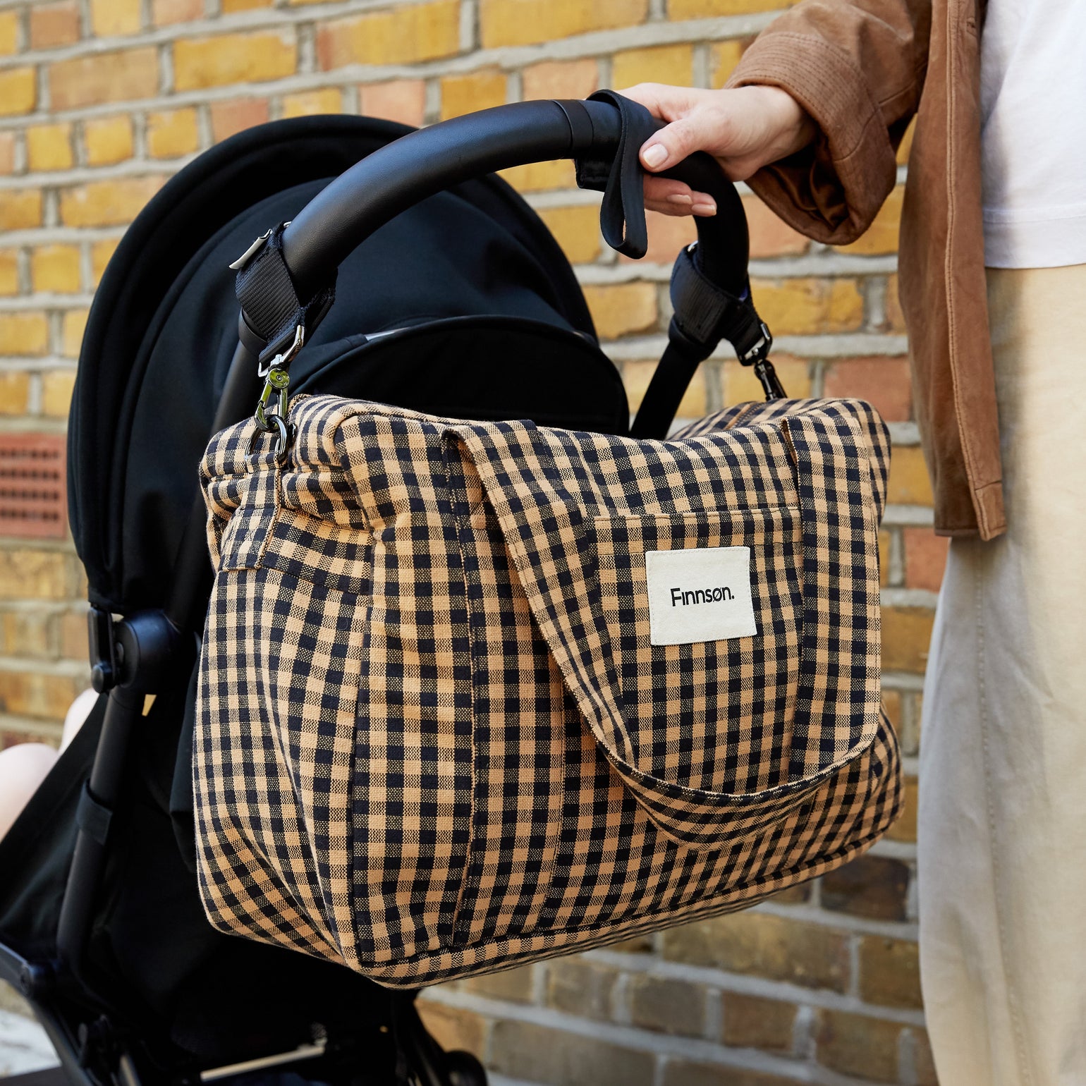 A person holds a black stroller next to the Sanna gingham check patterned bag, set against a yellow brick wall.