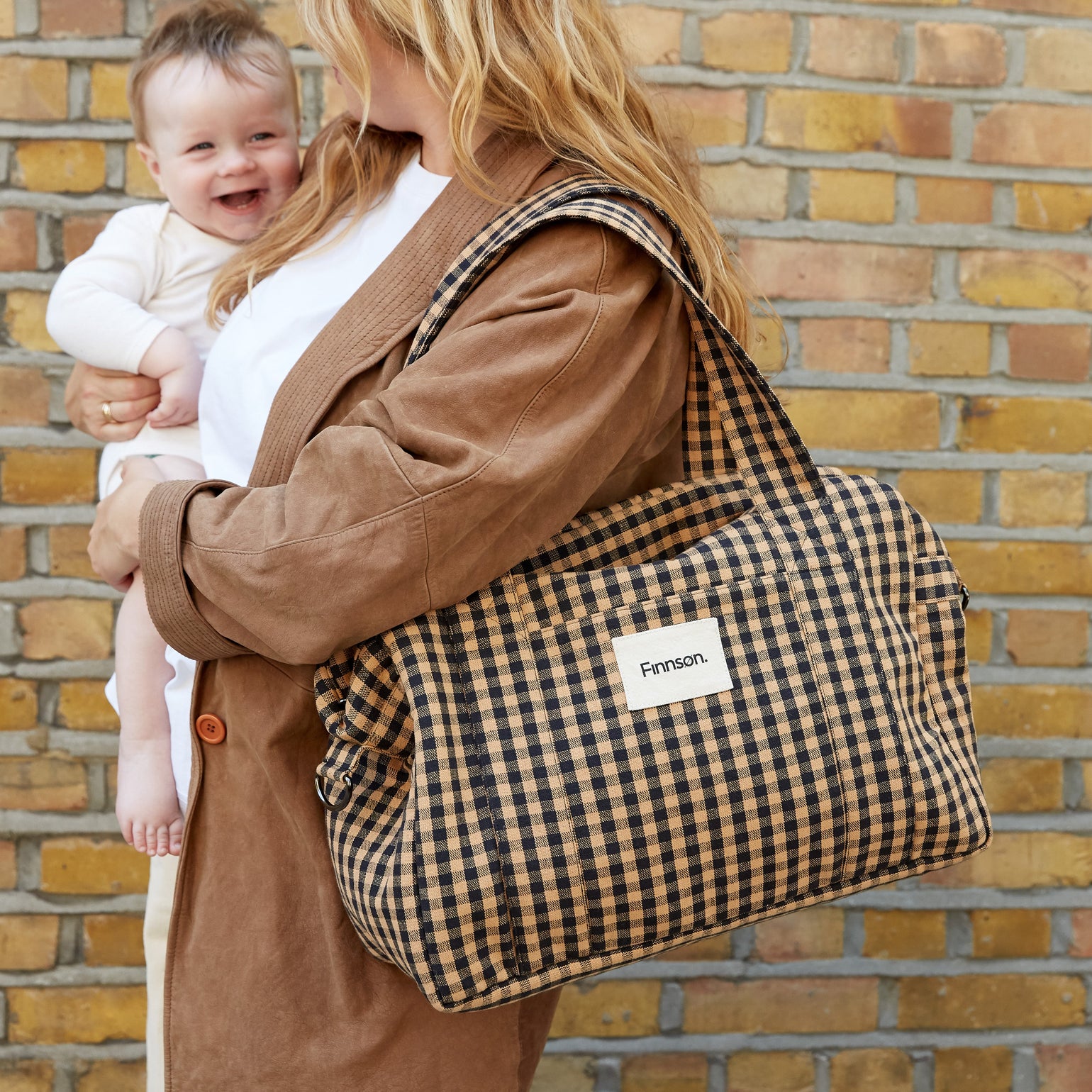 A woman carrying a baby while wearing the Sanna gingham check patterned bag, set against a brick wall background.