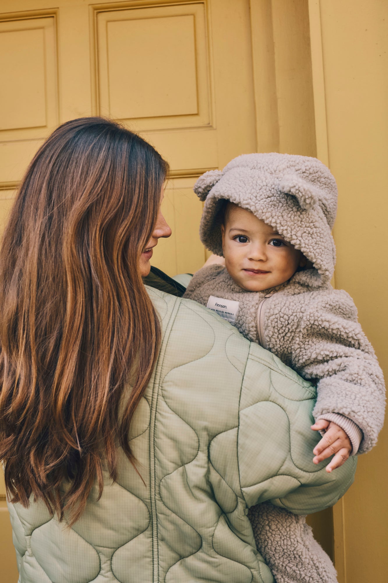 Woman holding a child in Alf borg beige eco pram suit against a yellow wall