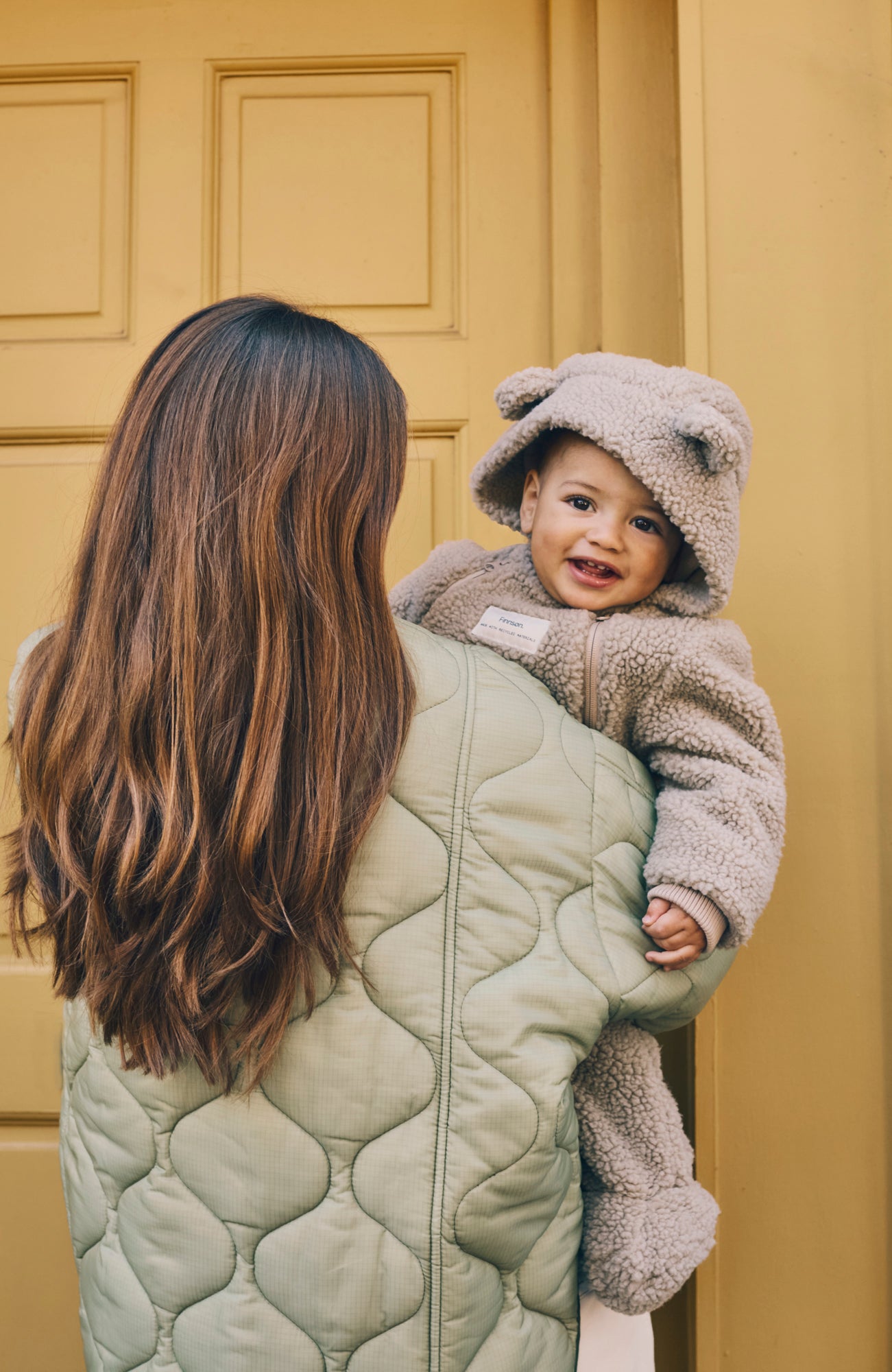 Woman holding a baby in a Alf borg beige eco pram suit against a yellow door.