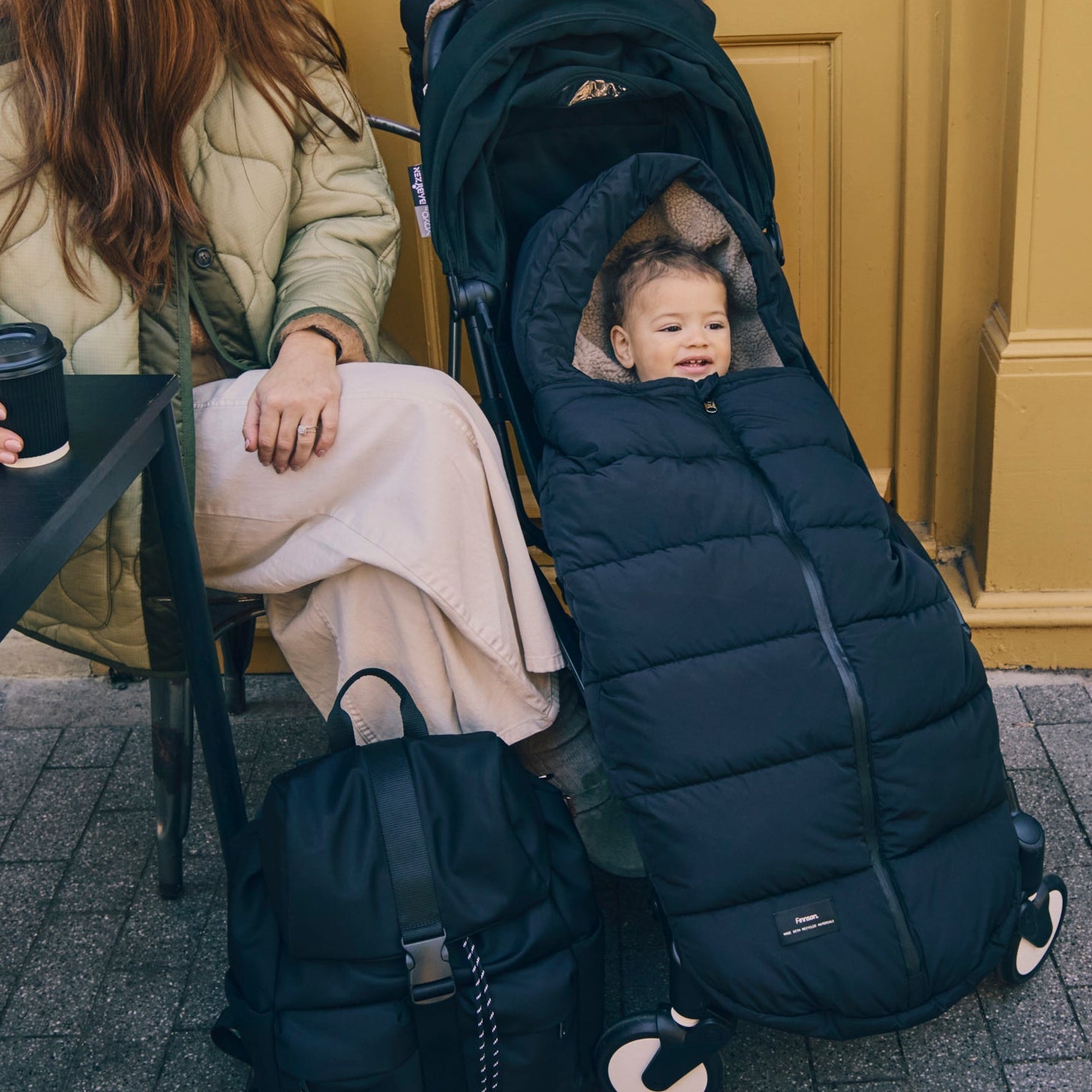 Child in a ALMA black footmuff with a women sitting nearby on a bench.
