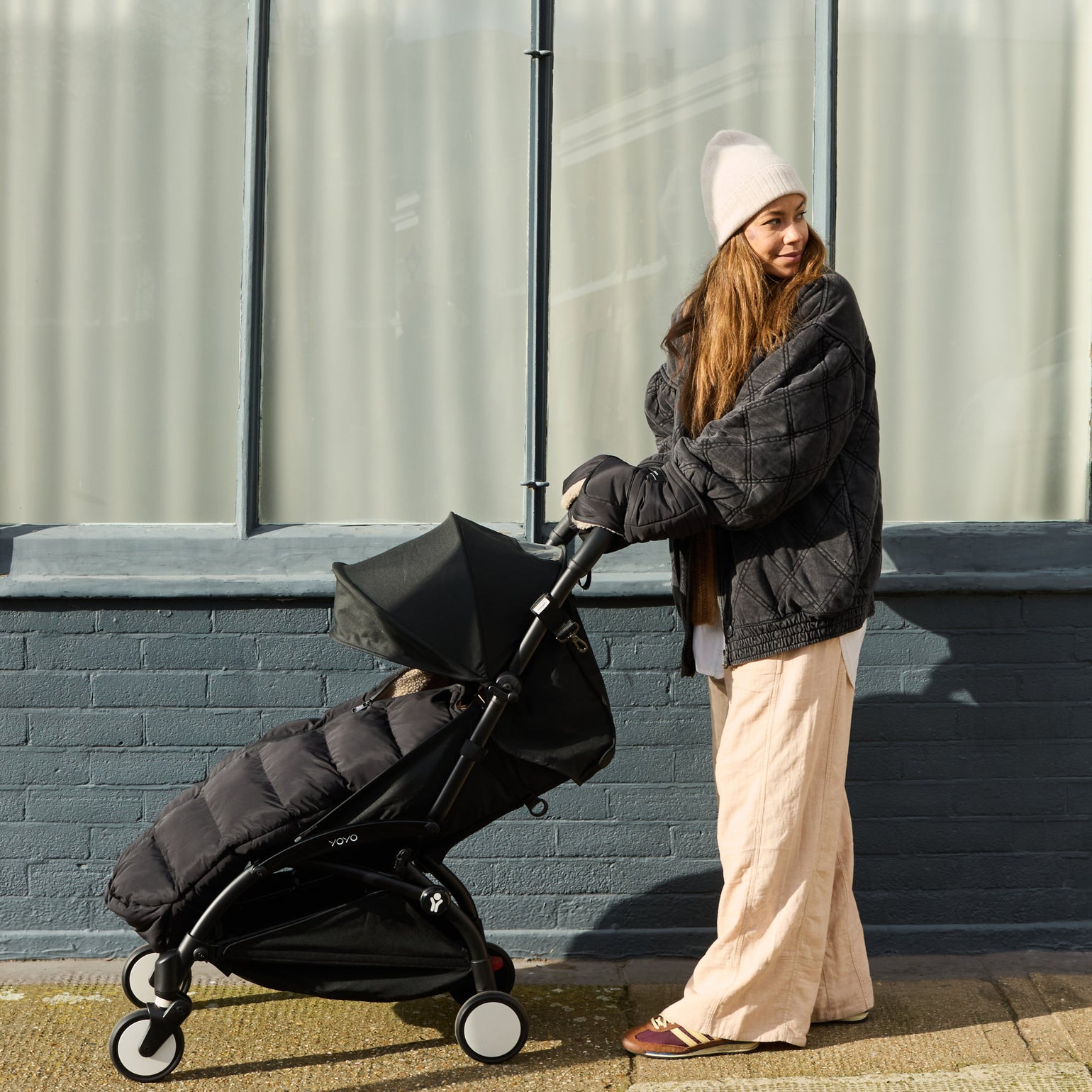 Woman pushing a stroller with the baby inside ALMA black footmuff against a building with large windows.