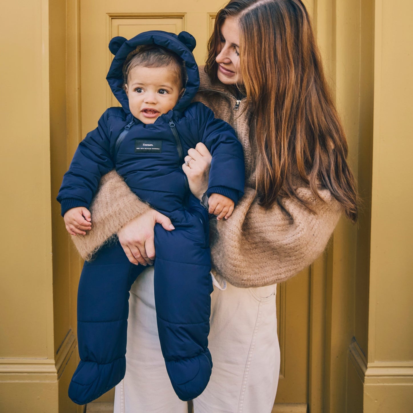Woman holding a baby wearing Anders navy snowsuit with bear ears against a yellow wall.