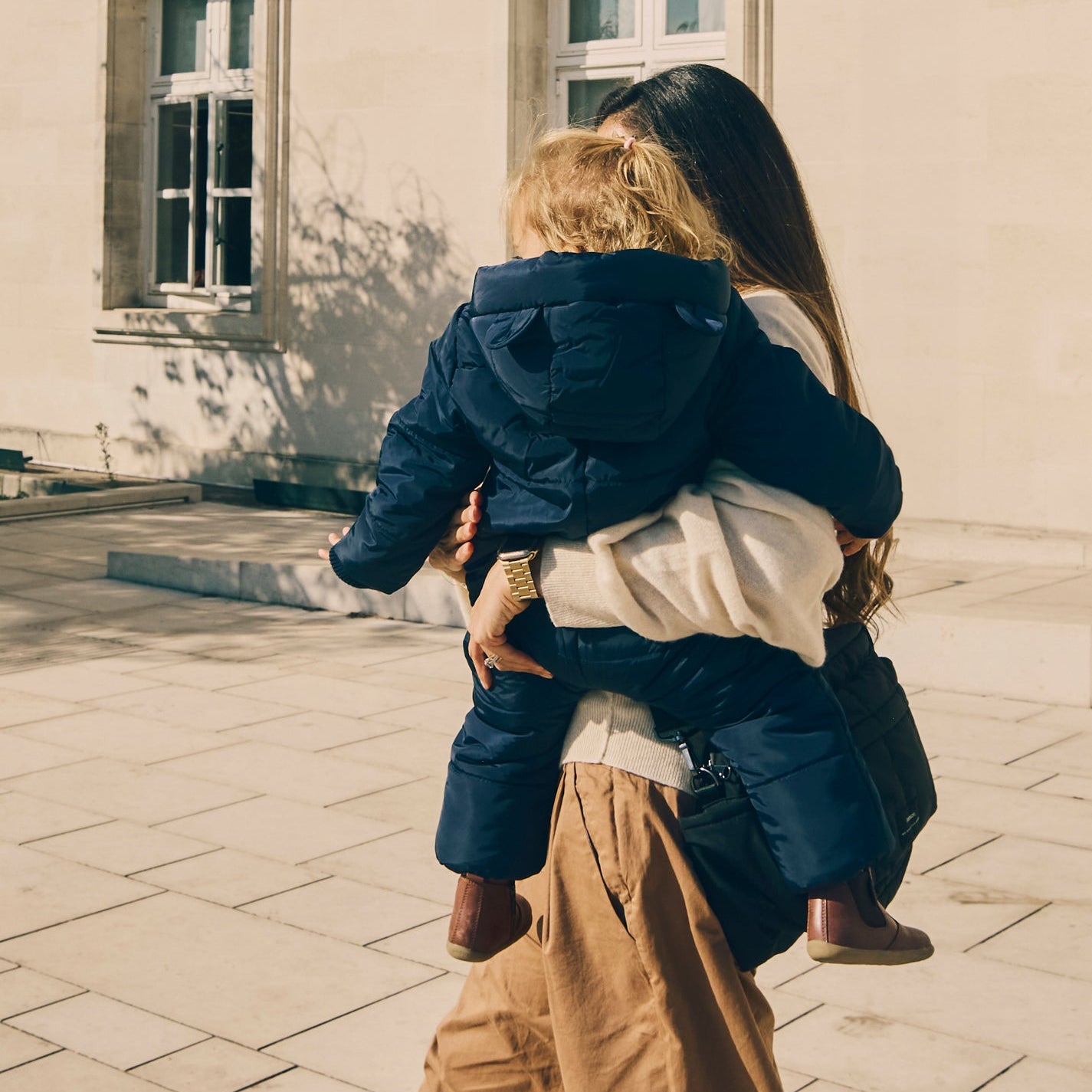 Woman holding a child wearing Anders navy snowsuit outdoors on a paved area with a building in the background