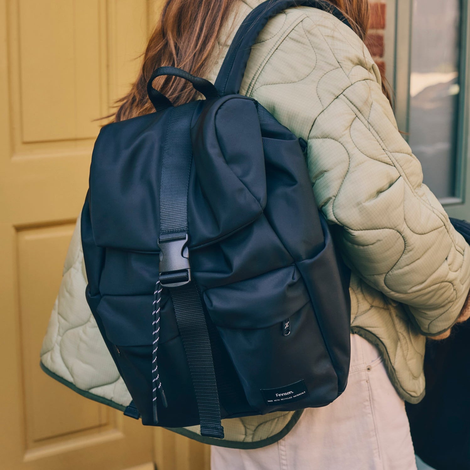 Person wearing the elise black changing backpack with a light green quilted jacket against a yellow door.