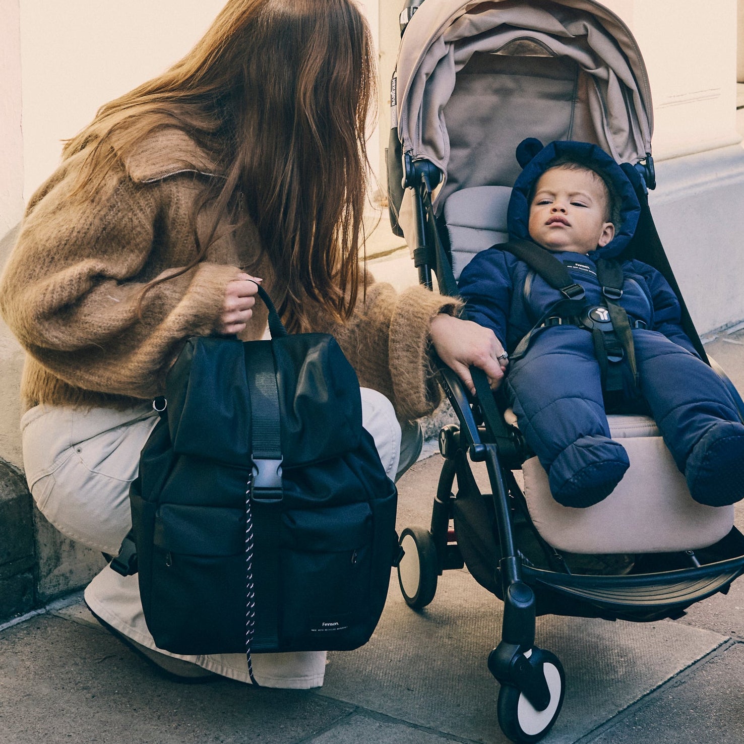 Woman with elise black changing backpack interacting with a child in a stroller on a sidewalk.