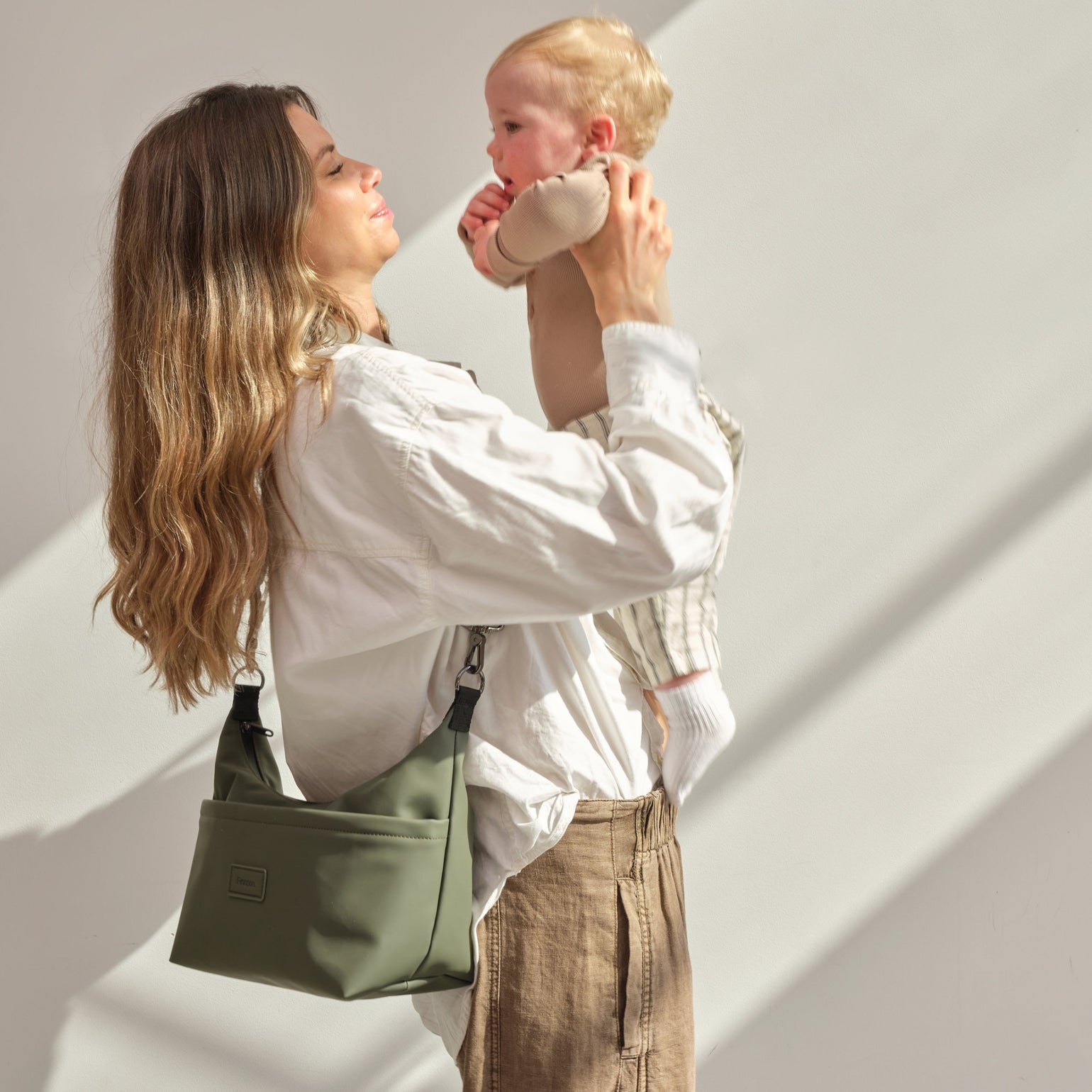 Woman holding a baby against a light gray wall with Fia khaki stroller organiser bag