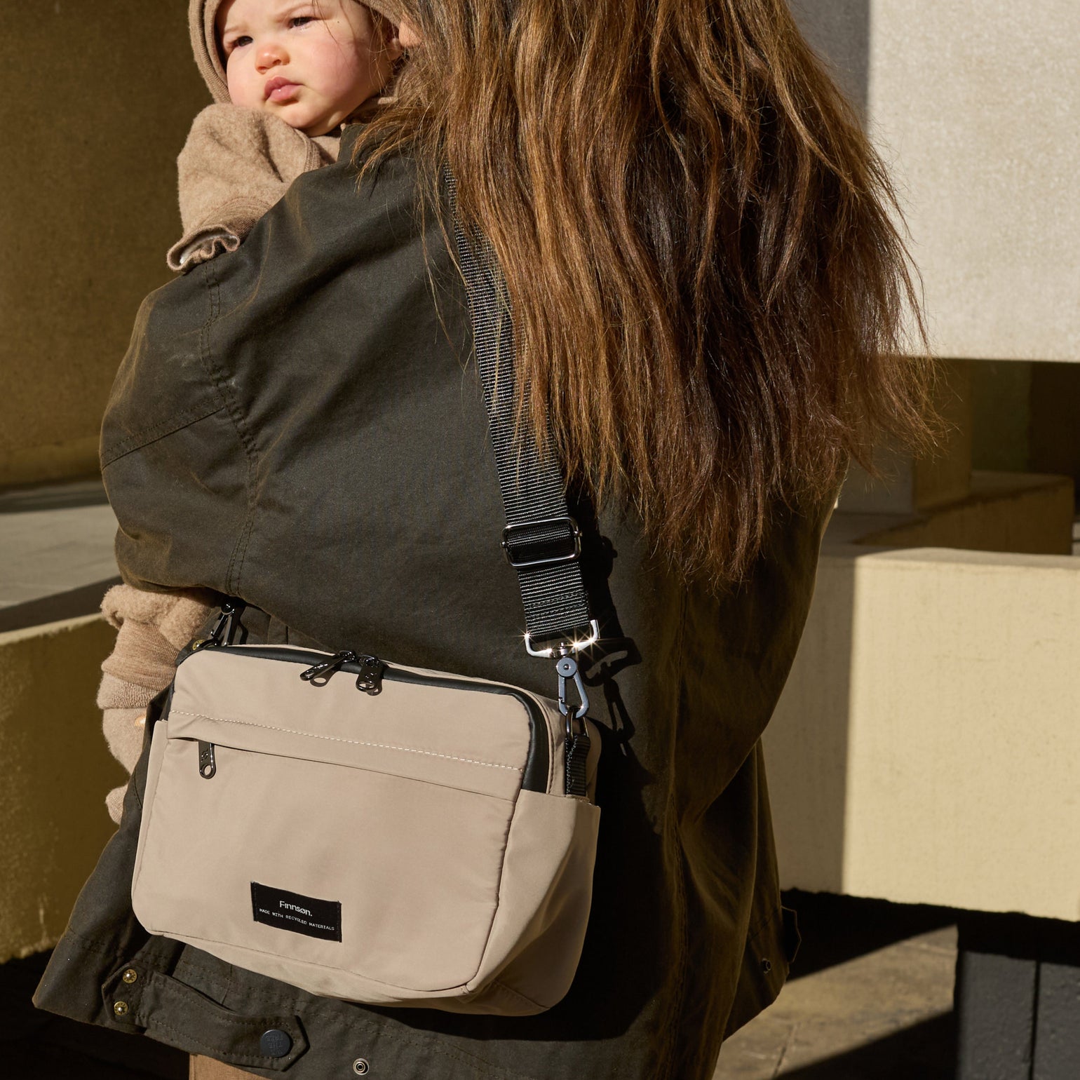 Frida taupe stroller organiser worn on shoulder whilst woman is carrying a baby