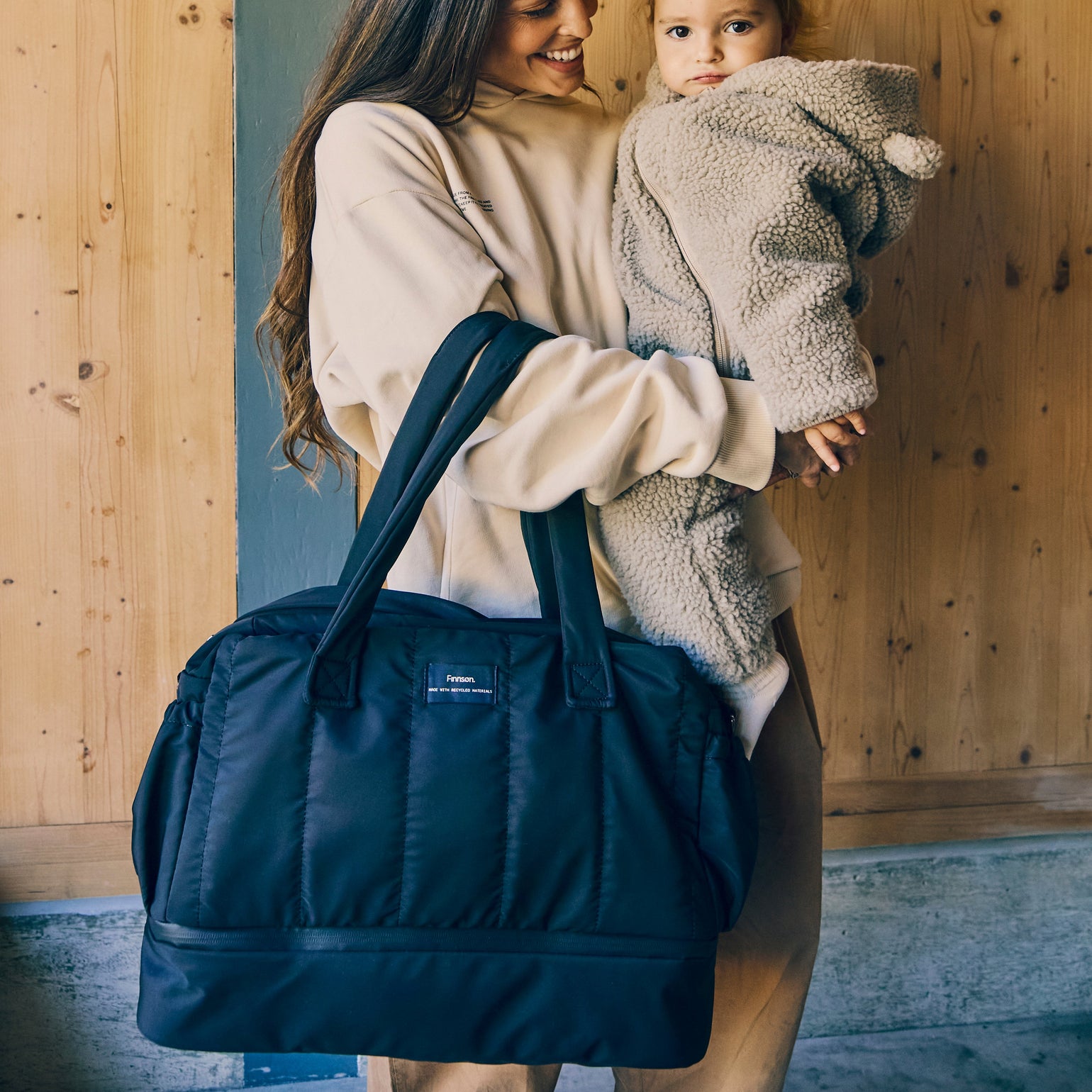 Woman holding a child and a Innes quilted black changing bag against a wooden wall.