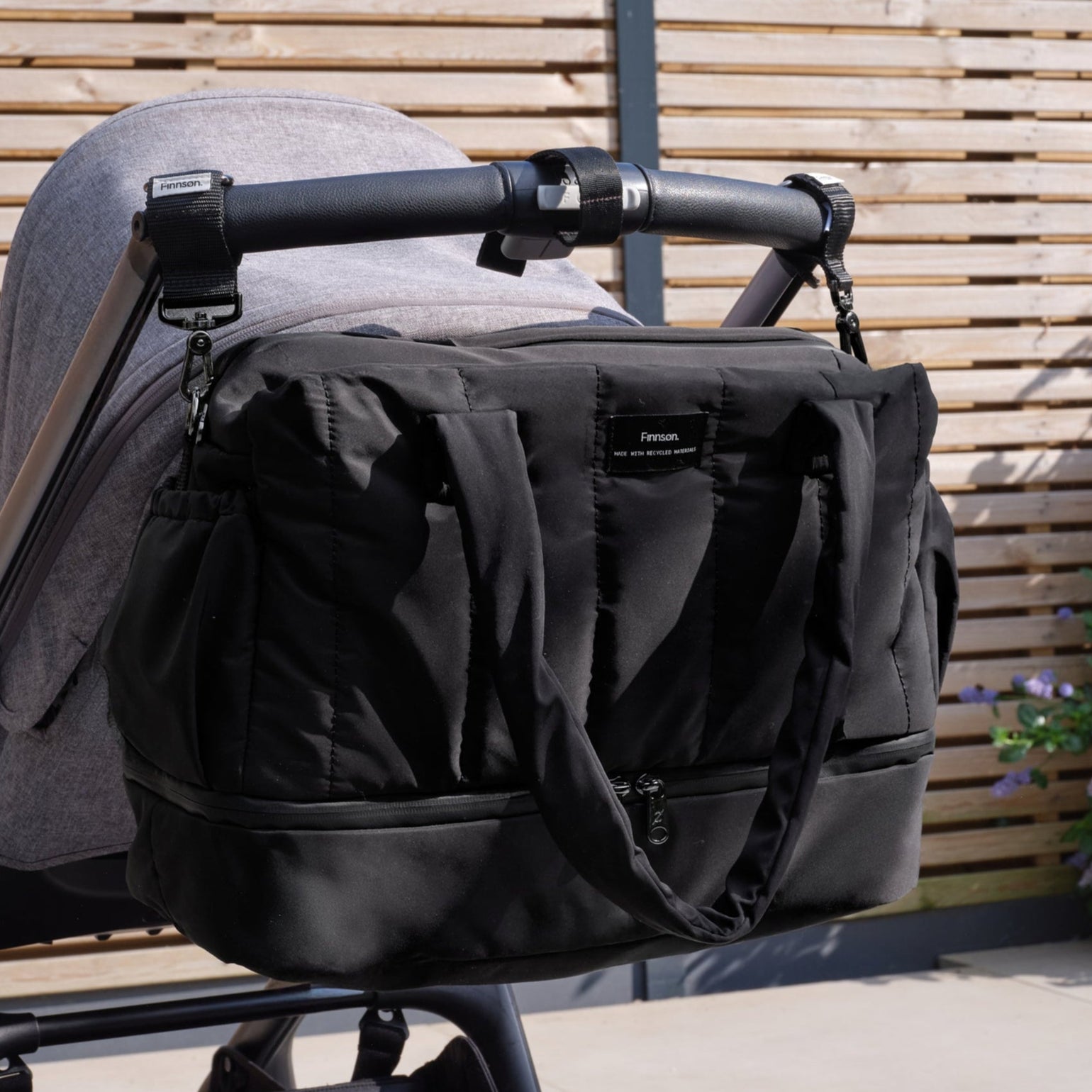 Innes quilted black changing bag attached to a stroller against a wooden fence background