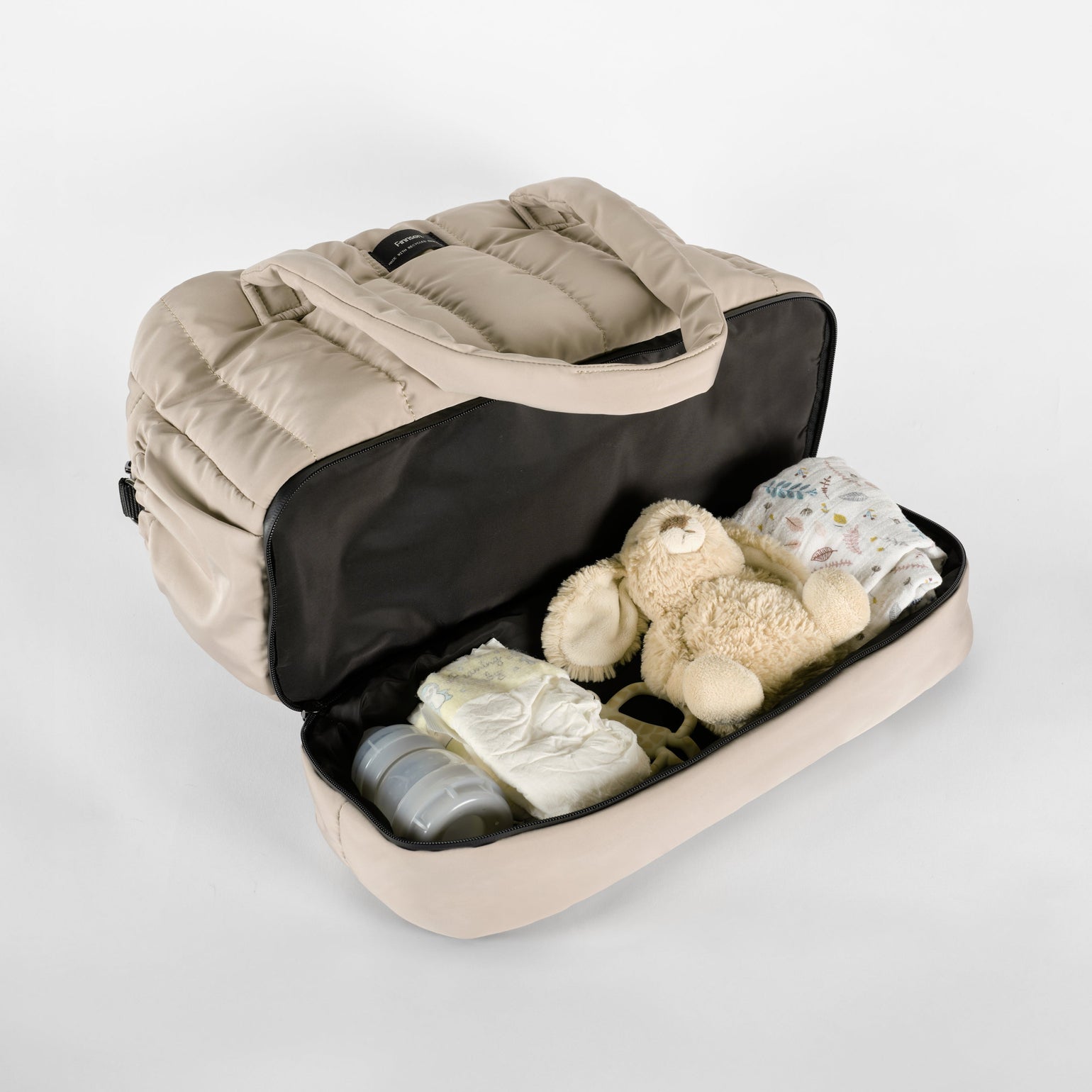 Open Innes Taupe quilted changing bag with a separate bottom storage compartment holding baby essentials, including nappies, bottles, clothing, and a comforter, showcasing its spacious, organised design for everyday parenting.