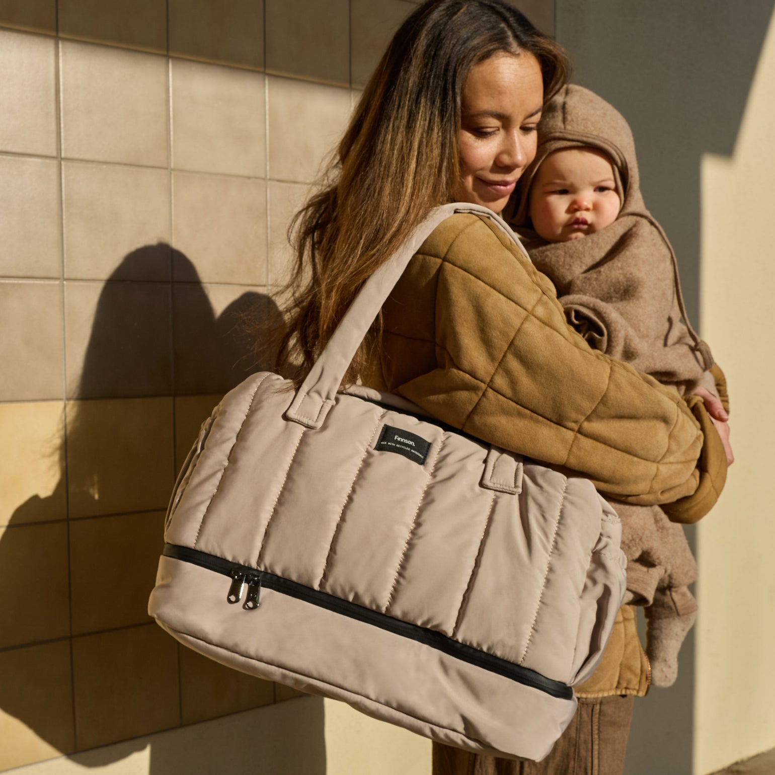 Parent carrying a baby while wearing the Innes Taupe quilted changing bag over the shoulder, showcasing its spacious quilted design, comfortable carry, and stylish practicality for everyday parenting.