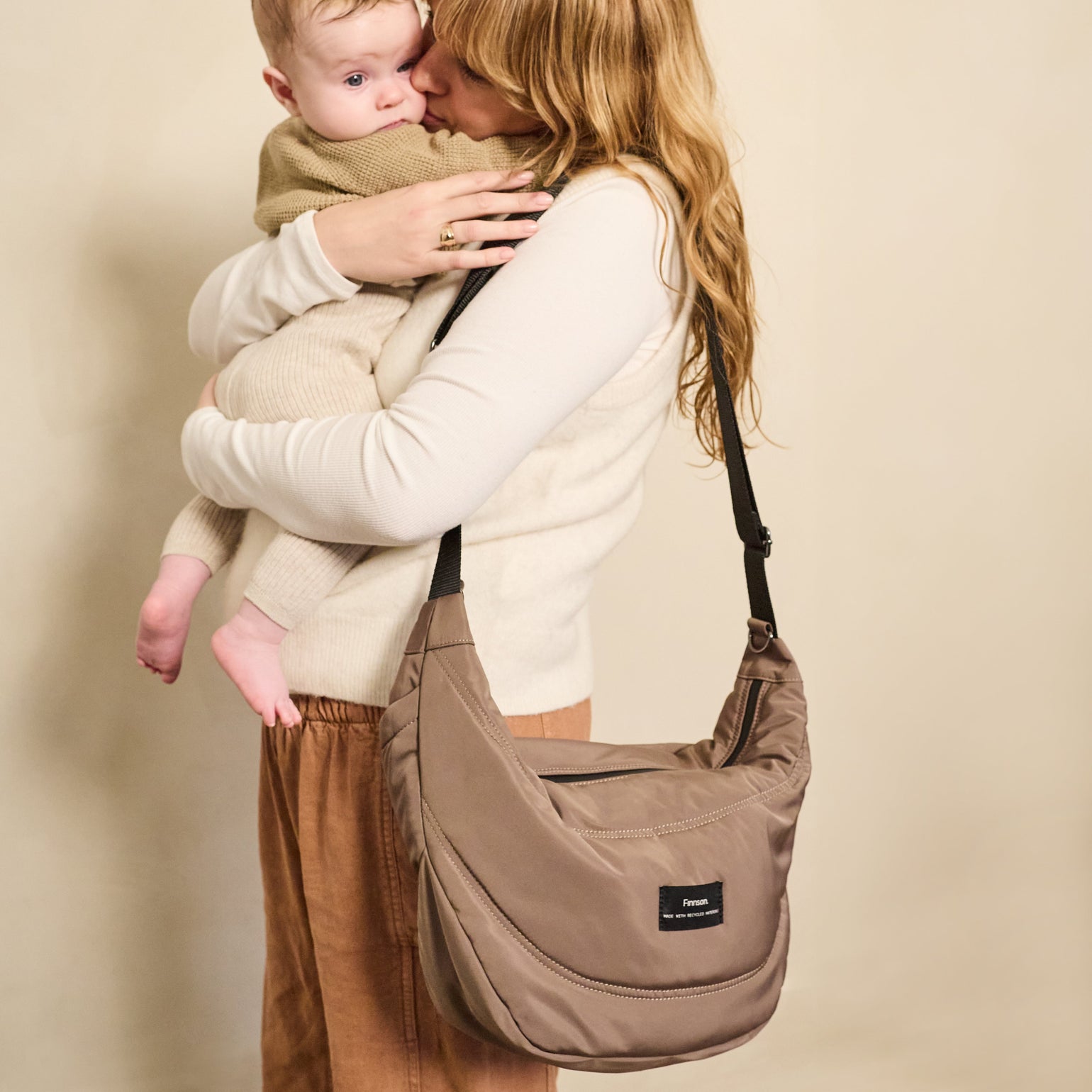 Woman holding a baby and carrying a finnson osma mocha stroller organiser against a beige background