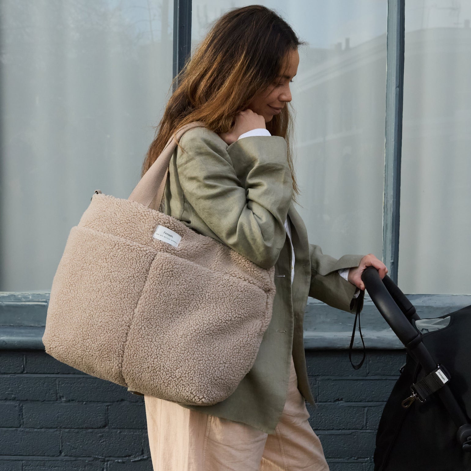 Woman holding a Sigrid mocha borg changing bag and pushing a stroller against a gray wall.