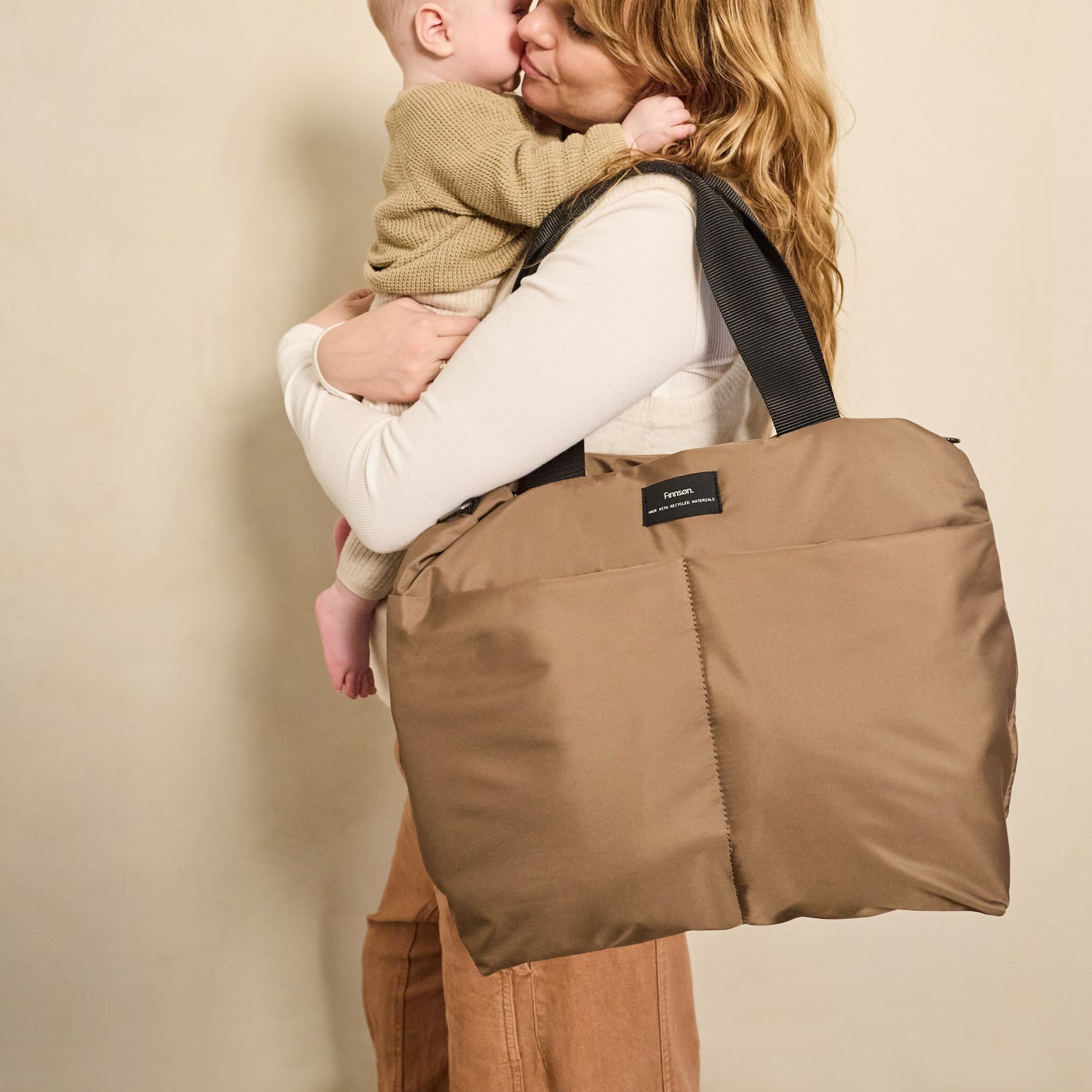 Woman carrying a child on her back with Sigrid Eco Stroller Organiser Bag in Mocha against a beige background