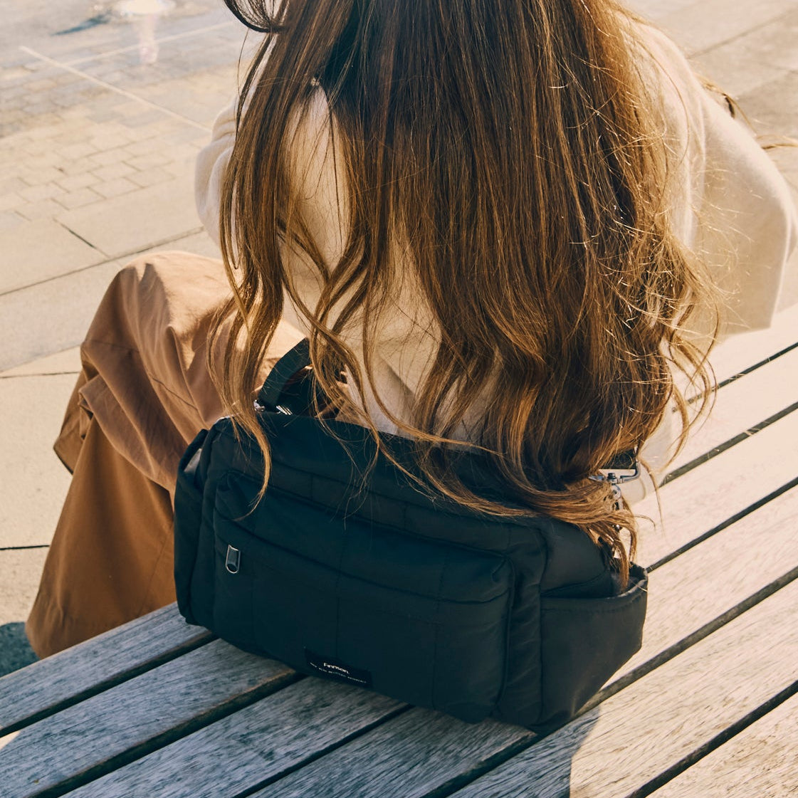 Person sitting on a bench with Freya eco stroller organiser black bag, wearing a beige coat and white shirt.