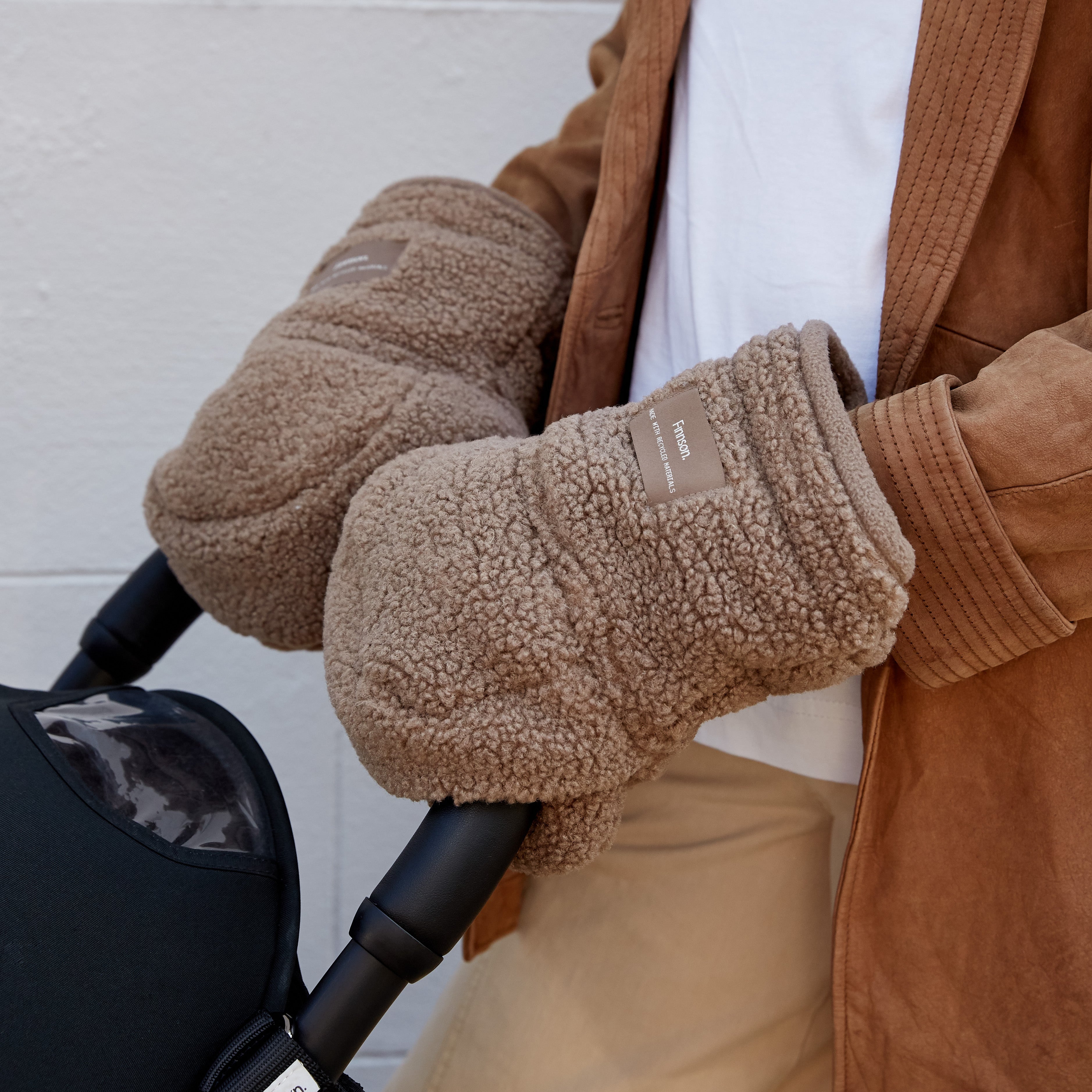 A close-up of a person wearing Astrid mocha borg universal pram stroller mittens, a brown corduroy jacket, and a white t-shirt, holding a black stroller handle against a white brick wall.