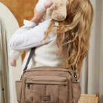 A woman carrying a baby over her shoulder while wearing a tan crossbody diaper bag. The woman is wearing a cream sweater and brown pants, and the baby is holding a beige teddy bear.