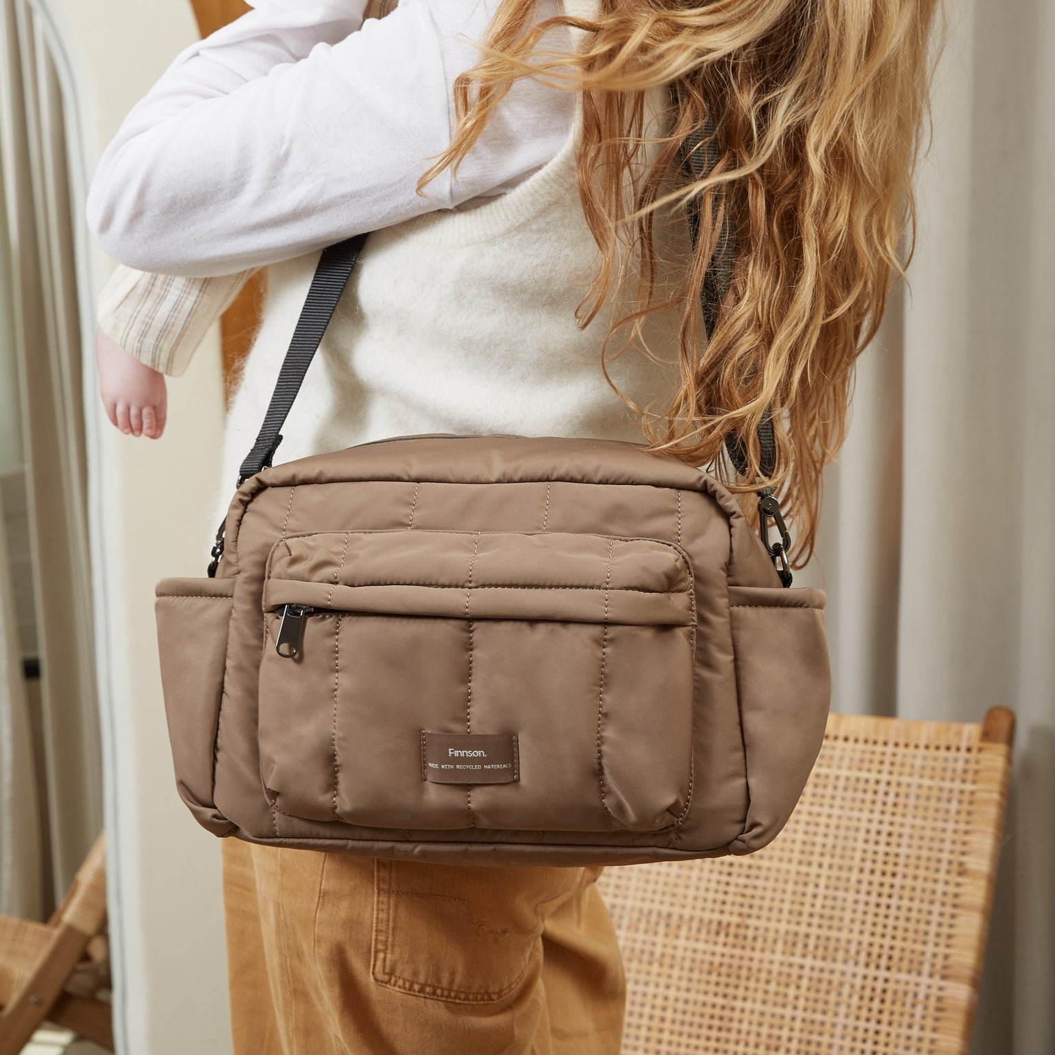 A woman carrying a baby over her shoulder while wearing a tan crossbody diaper bag. The woman is wearing a cream sweater and brown pants, and the baby is holding a beige teddy bear.