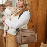 A woman wearing a white long-sleeve top and tan pants carries a baby in a cream baby carrier while wearing a mocha brown shoulder bag. She is standing in front of a wooden wall mirror.
