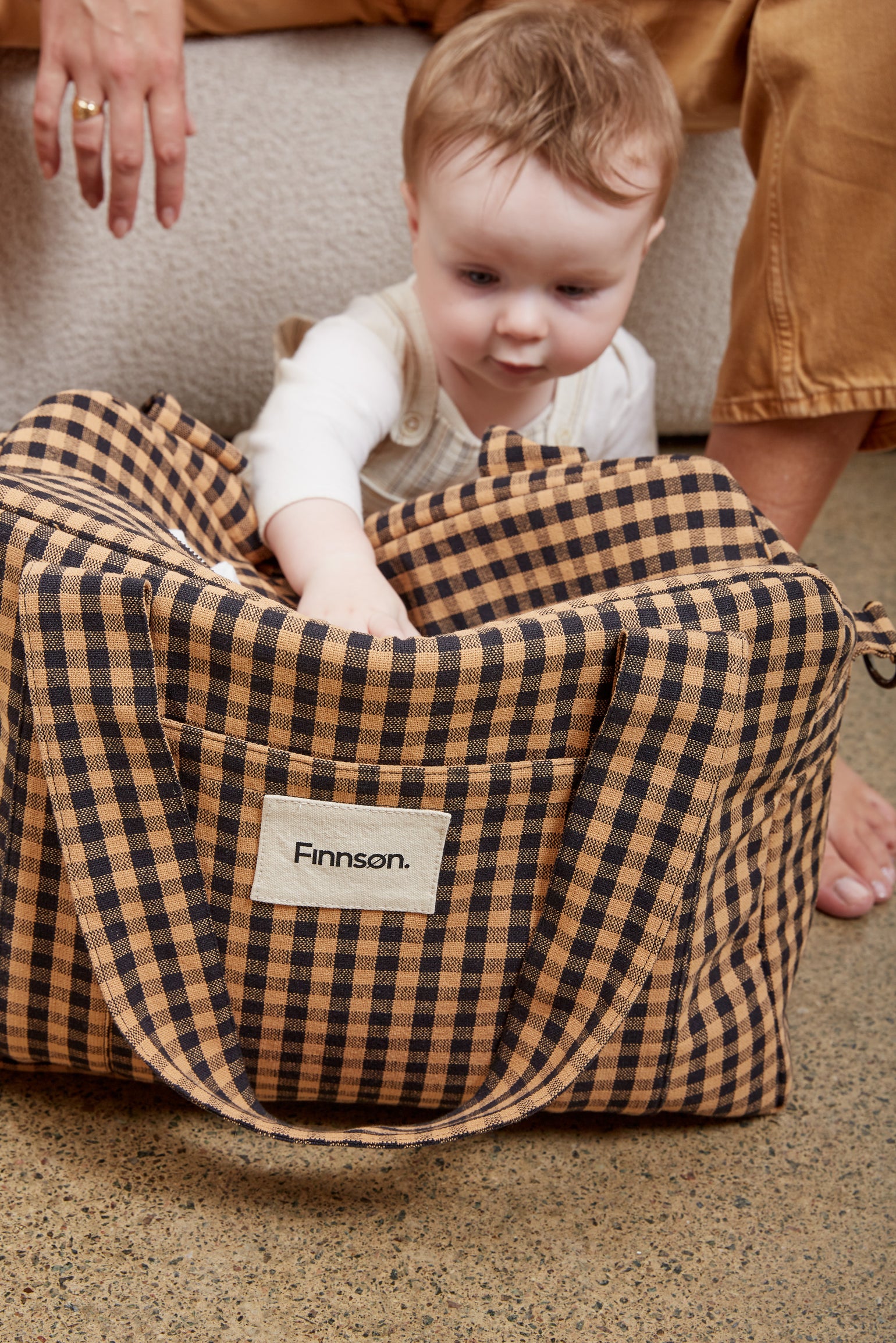 A toddler sits beside the Sanna gingham check patterned bag, with an adult's hand and legs visible in the background. The bag features a front pocket with a white label that reads 'Finnson'.