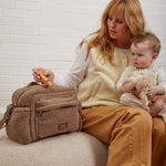 A woman sits on a beige upholstered bench holding a baby, with freya mocha borg bag beside her. The woman wears a cream vest over a white long-sleeve top and mustard-brown pants.
