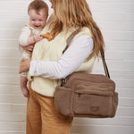 A woman holding a baby stands against a white brick wall, wearing a freya mocha borg bag. The woman wears a cream vest over a white long-sleeve shirt and tan pants.