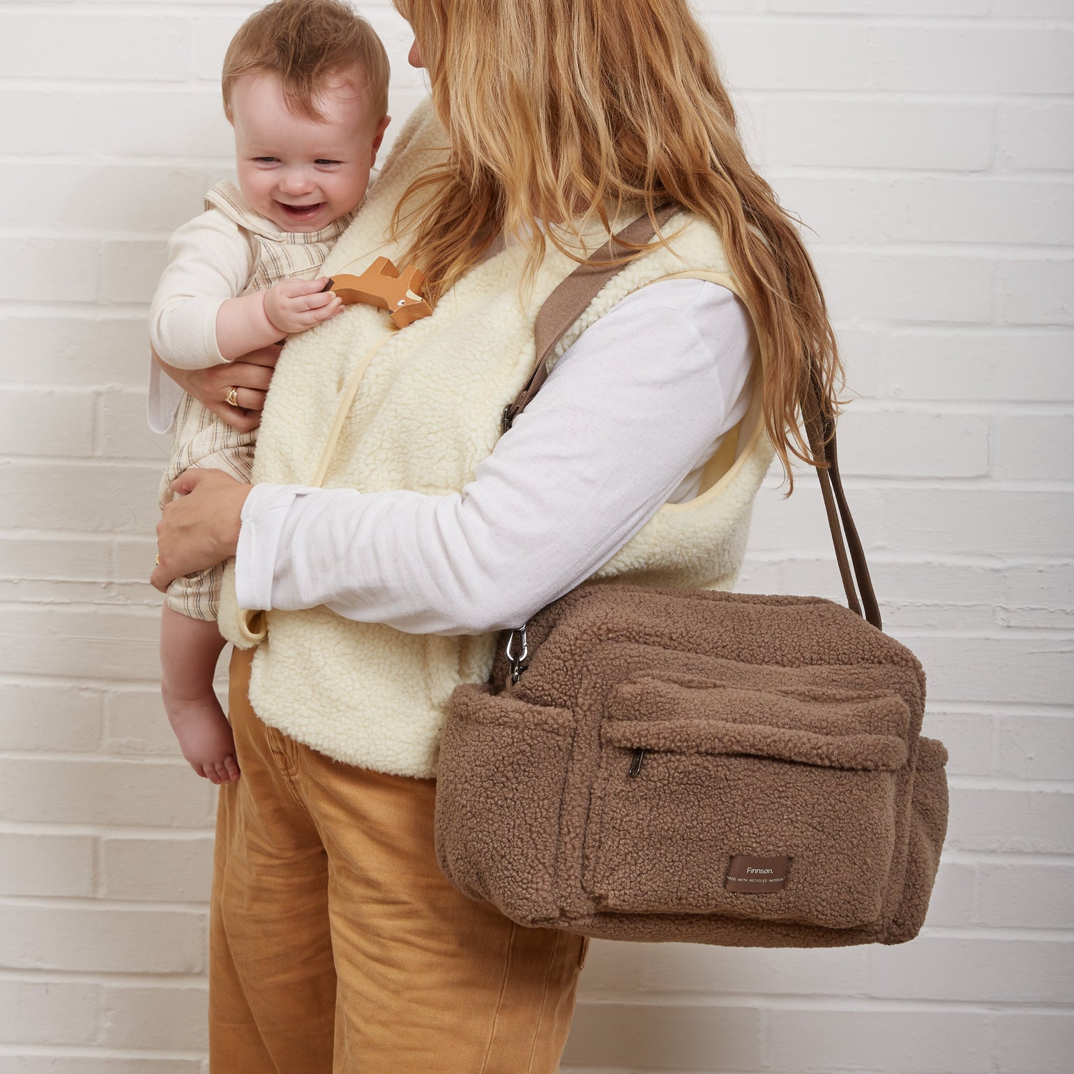 A woman holding a baby stands against a white brick wall, wearing a freya mocha borg bag. The woman wears a cream vest over a white long-sleeve shirt and tan pants.