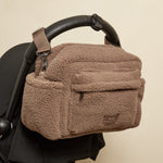 A freya mocha borg stroller bag with a small logo patch, hanging from a black stroller handle against a plain beige wall.