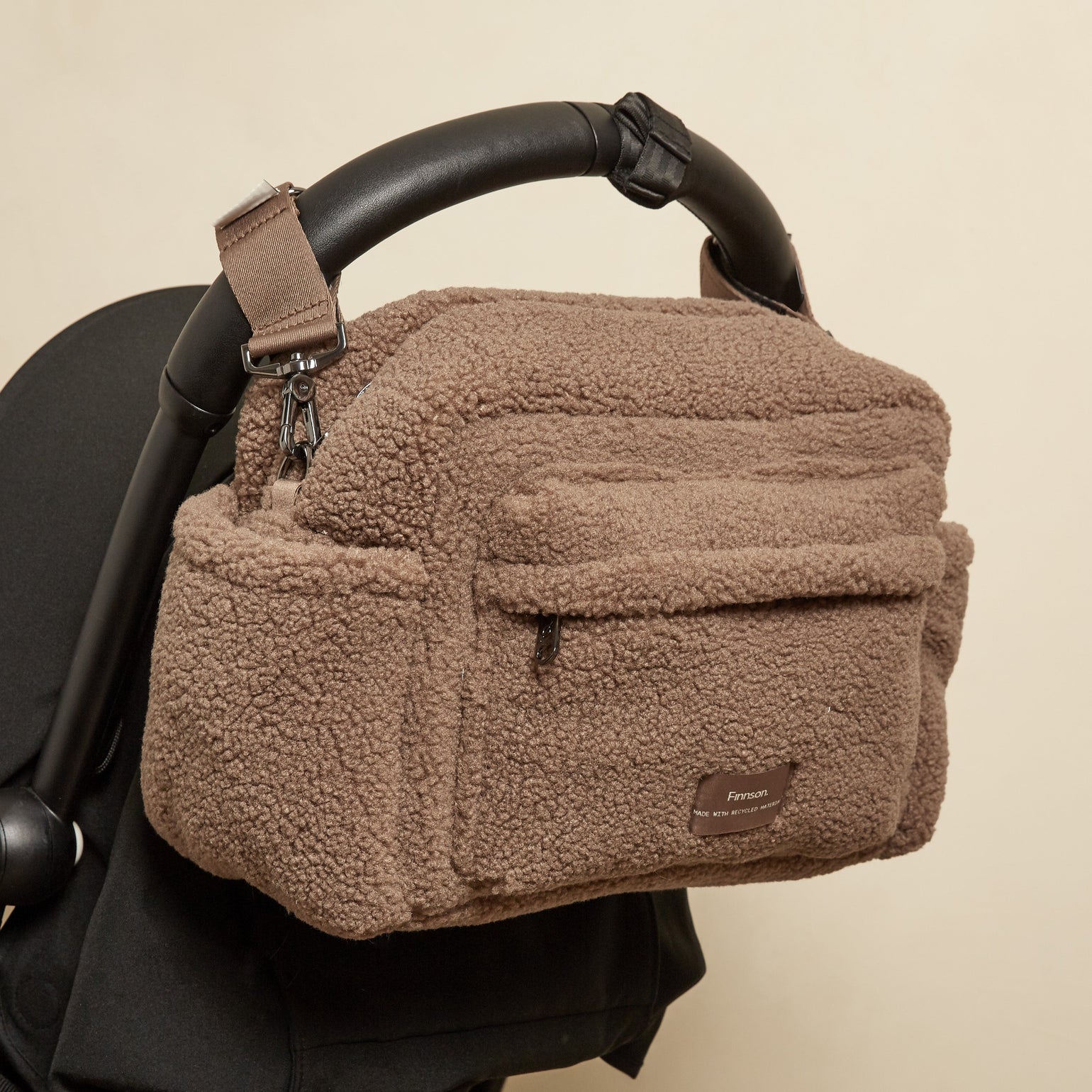 A freya mocha borg stroller bag with a small logo patch, hanging from a black stroller handle against a plain beige wall.
