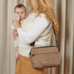 A woman holding a baby wearing a mocha crossbody diaper bag with a black strap, set against a neutral beige curtain background.