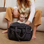 A woman and a baby sit on a beige pouf in a bright room, with the Finnson Iris changing bag placed in front of them on the carpet.