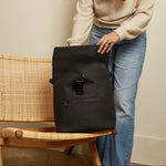 A woman in a beige sweater and blue jeans stands beside a woven chair holding the Finnson rolltop black backpack. The backpack has a front zip pocket with a strap closure and a small rectangular logo patch near the bottom.