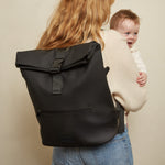 A woman with long blonde hair, seen from behind, carries a baby in a cream knit carrier while wearing the Finnson Viggo black roll-top backpack. She is wearing blue jeans against a plain beige studio background.