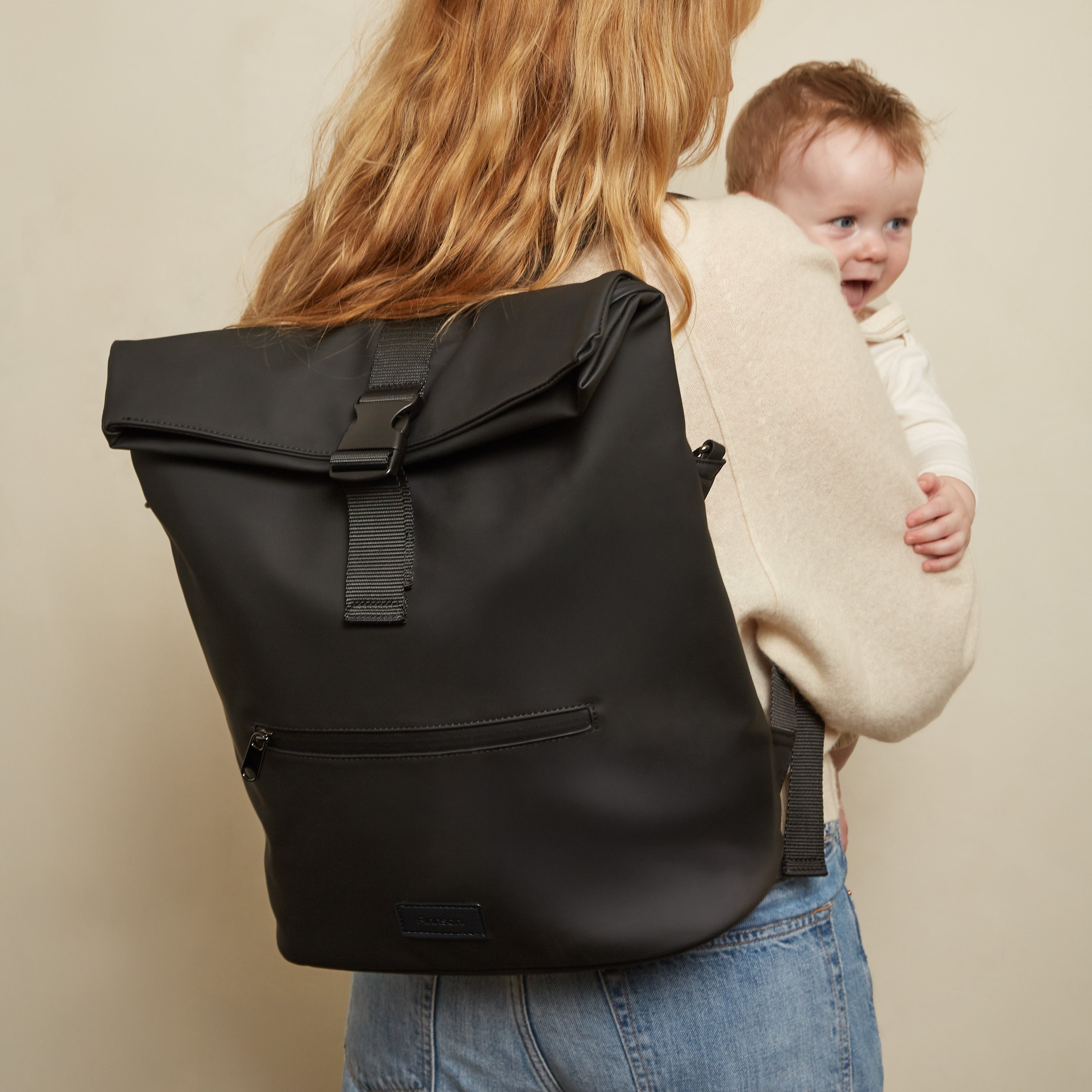 A woman with long blonde hair, seen from behind, carries a baby in a cream knit carrier while wearing the Finnson Viggo black roll-top backpack. She is wearing blue jeans against a plain beige studio background.