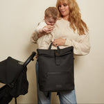 A woman holding a baby stands next to a black stroller and carries the Finnson viggo rolltop black backpack changing bag with top handles and a front zip pocket, against a plain beige wall.