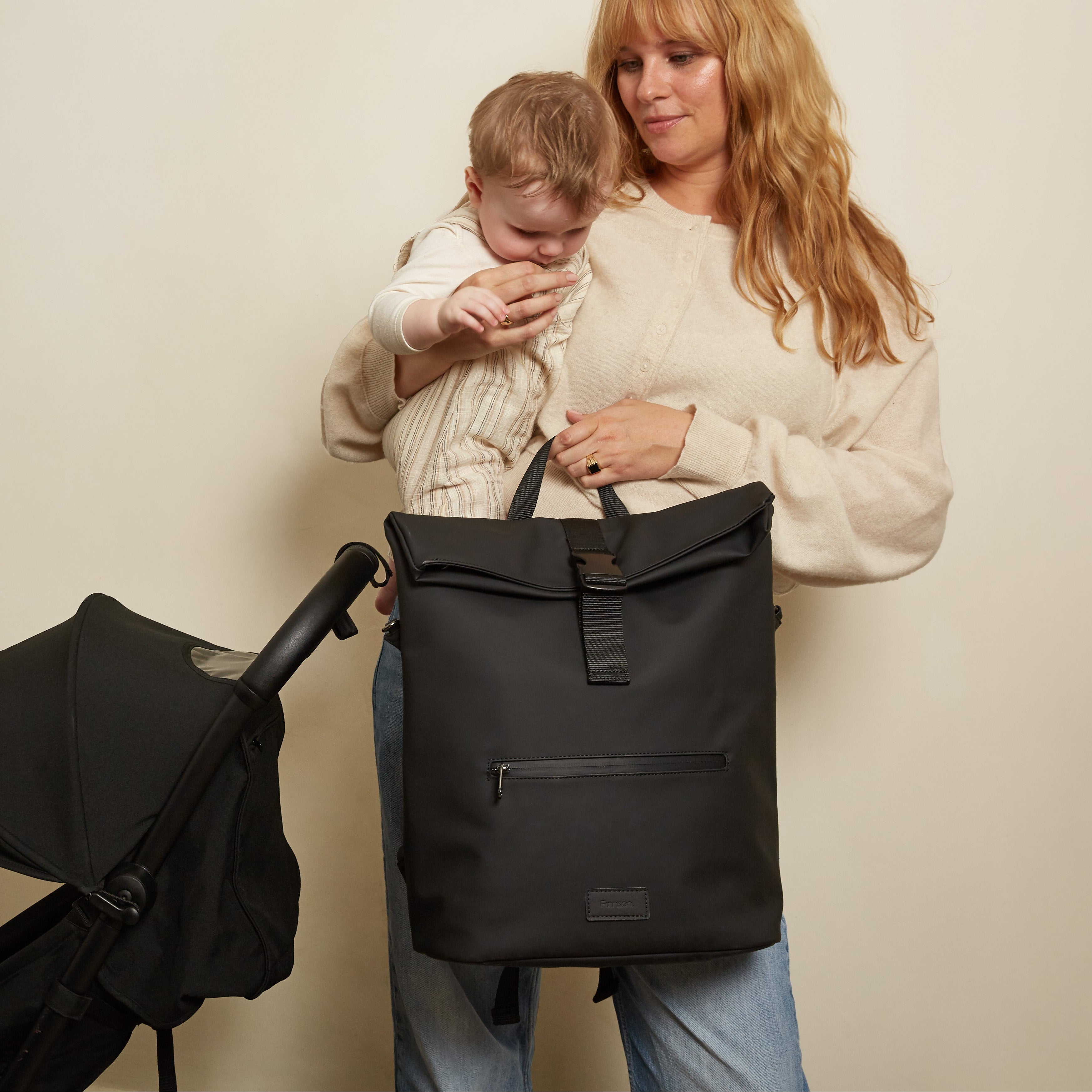 A woman holding a baby stands next to a black stroller and carries the Finnson viggo rolltop black backpack changing bag with top handles and a front zip pocket, against a plain beige wall.