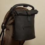 The Finnson Viggo black roll-top diaper bag with a front zip pocket and buckle strap, shown hanging from a brown stroller against a plain beige wall.