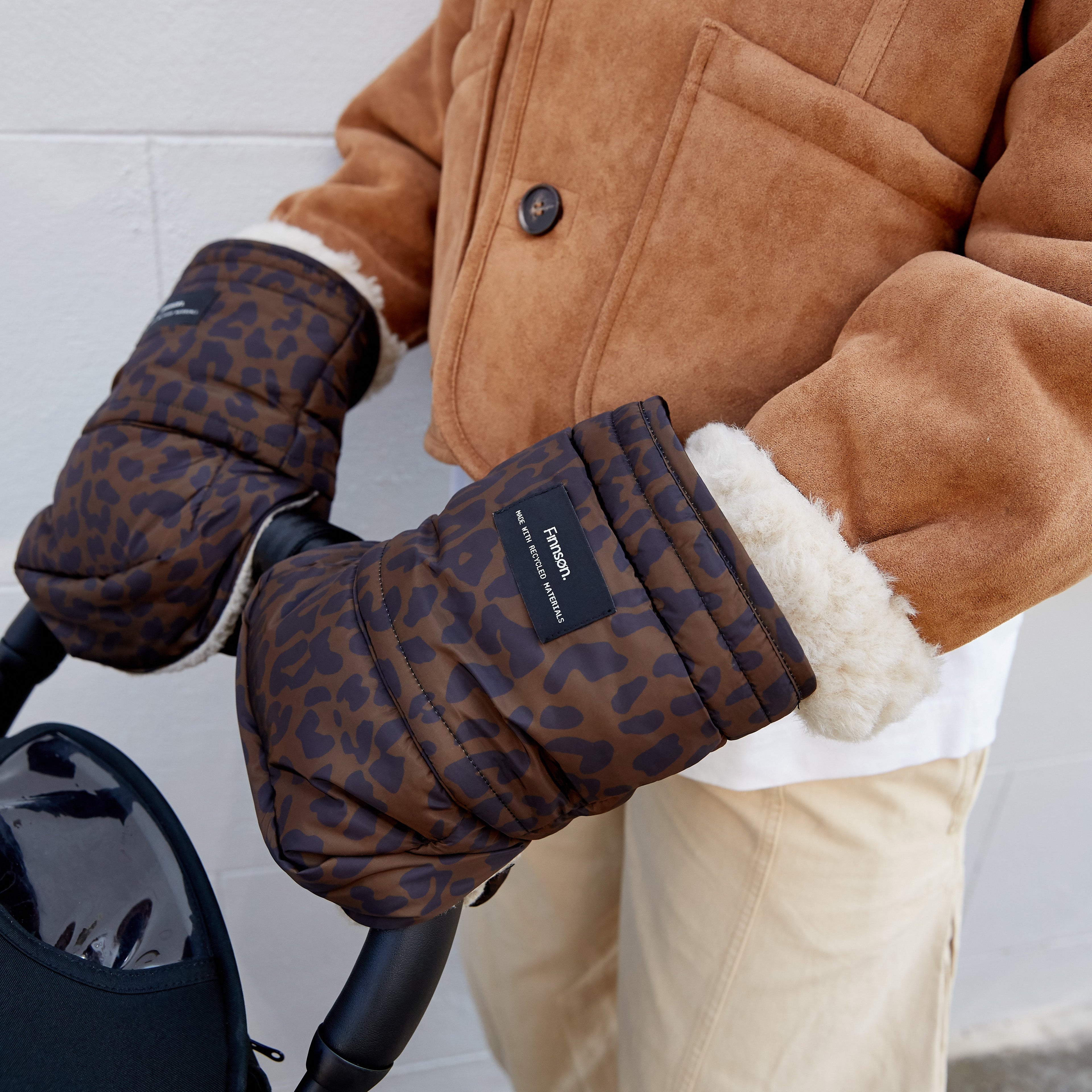 Astrid chocolate brown leopard stroller mittens attached to stroller. Woman is wearing with a tan suede jacket and beige pants, holding a stroller handle.