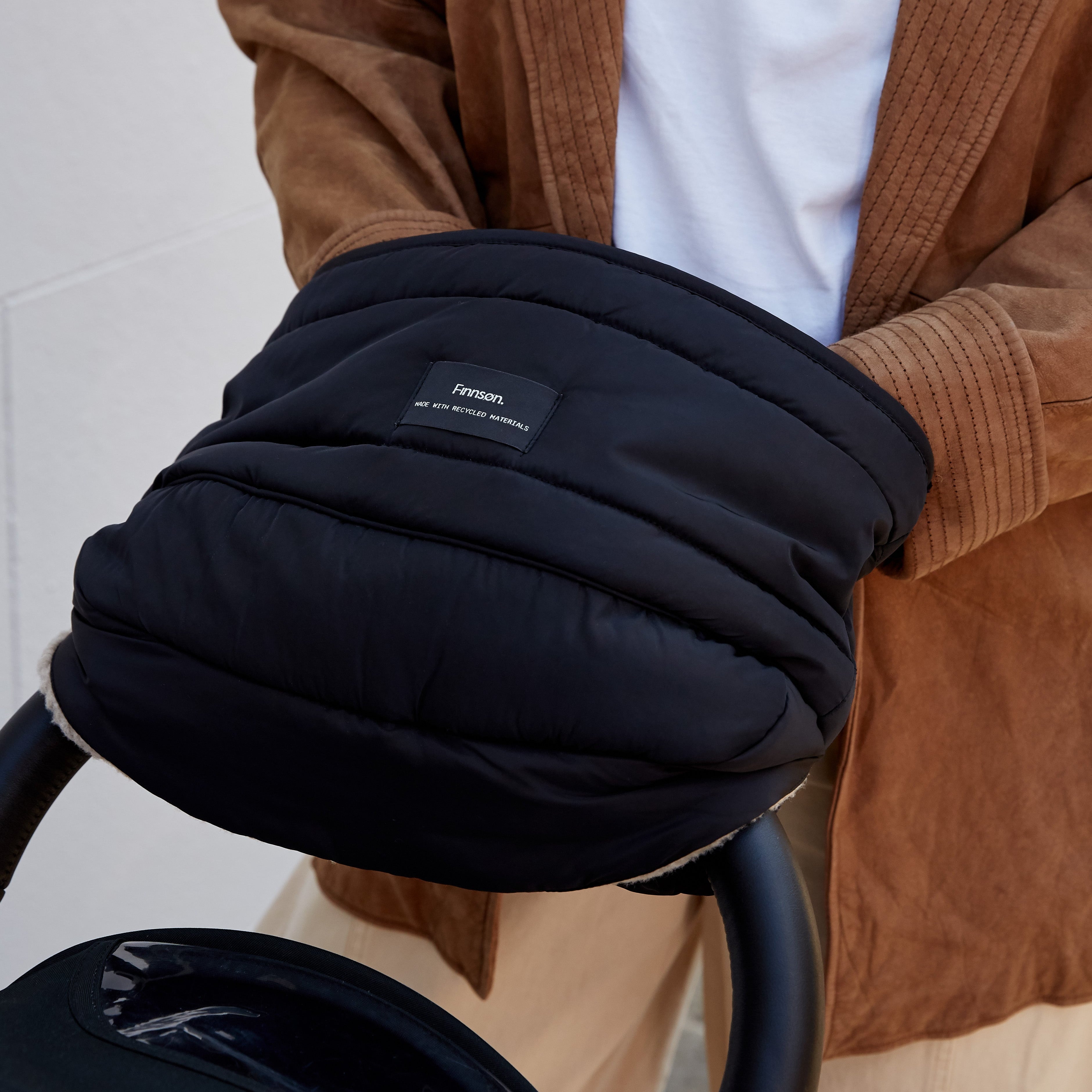 A close-up of a person wearing a brown leather jacket over a white shirt, holding the finnson Agnar black padded stroller mitten or stroller accessory attached to a stroller.
