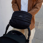 A woman in a brown leather jacket and white top pushes a black stroller while wearing the agnar black padded stroller mitten. The stroller has a small white logo on the bag.