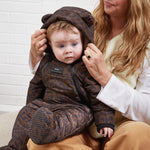 A baby wearing the Anders a brown and black leopard-print padded snowsuit with a bear-ear hood, sitting on a woman's lap against a white brick wall.