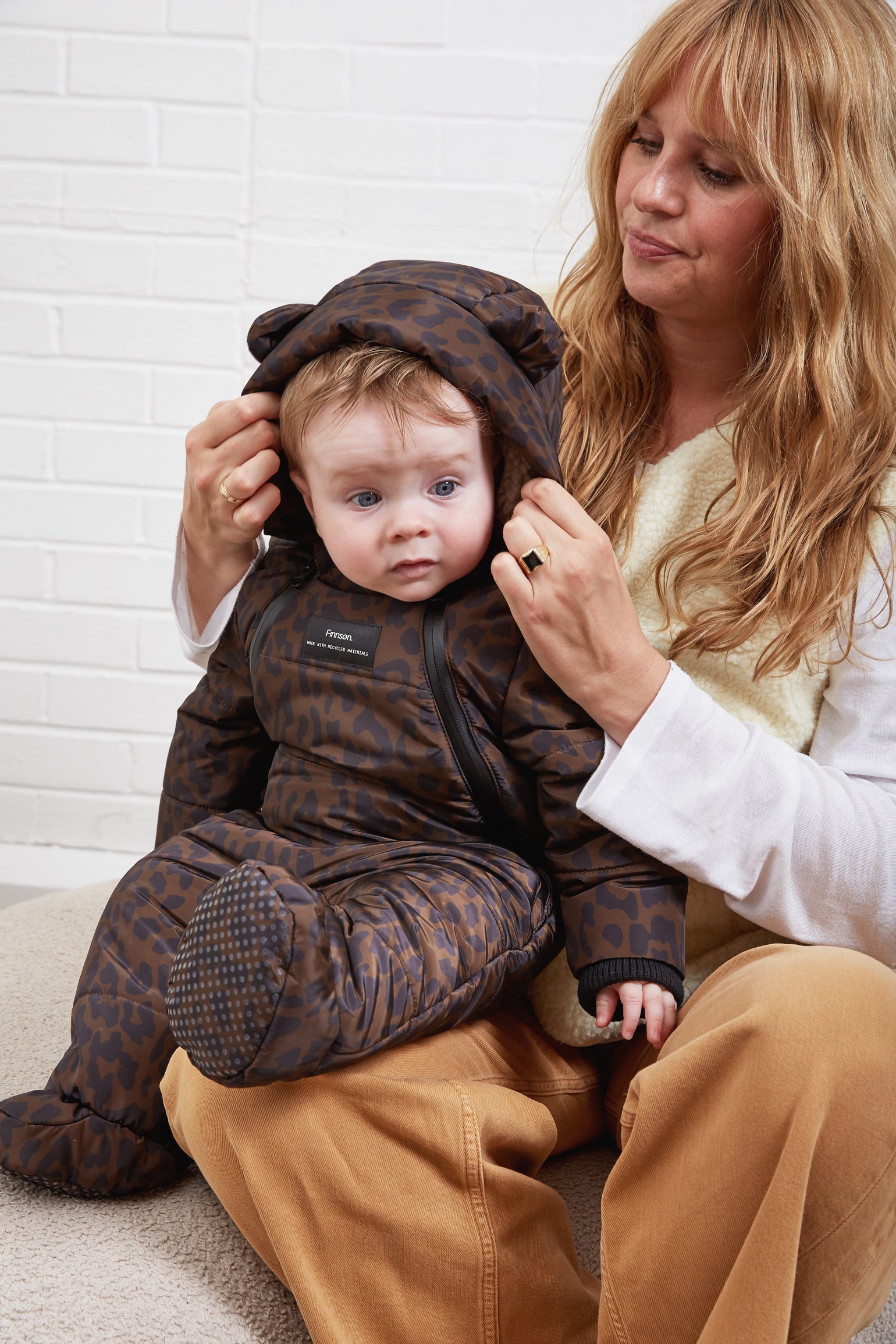 A baby wearing the Anders a brown and black leopard-print padded snowsuit with a bear-ear hood, sitting on a woman's lap against a white brick wall.