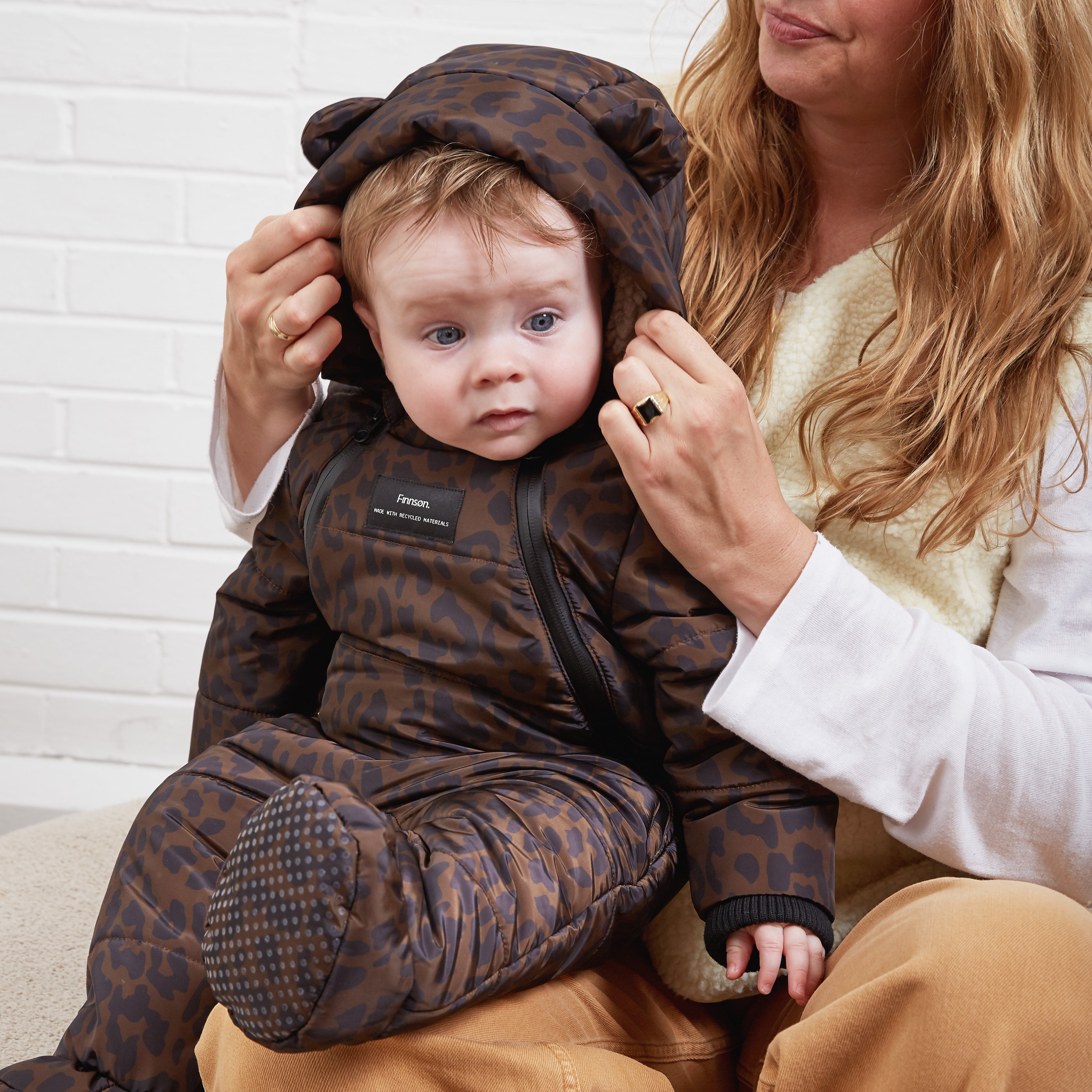 A baby wearing the Anders a brown and black leopard-print padded snowsuit with a bear-ear hood, sitting on a woman's lap against a white brick wall.