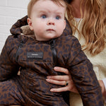 A baby wearing anders choco leopard print snow suit, held by a woman against a white brick wall background.