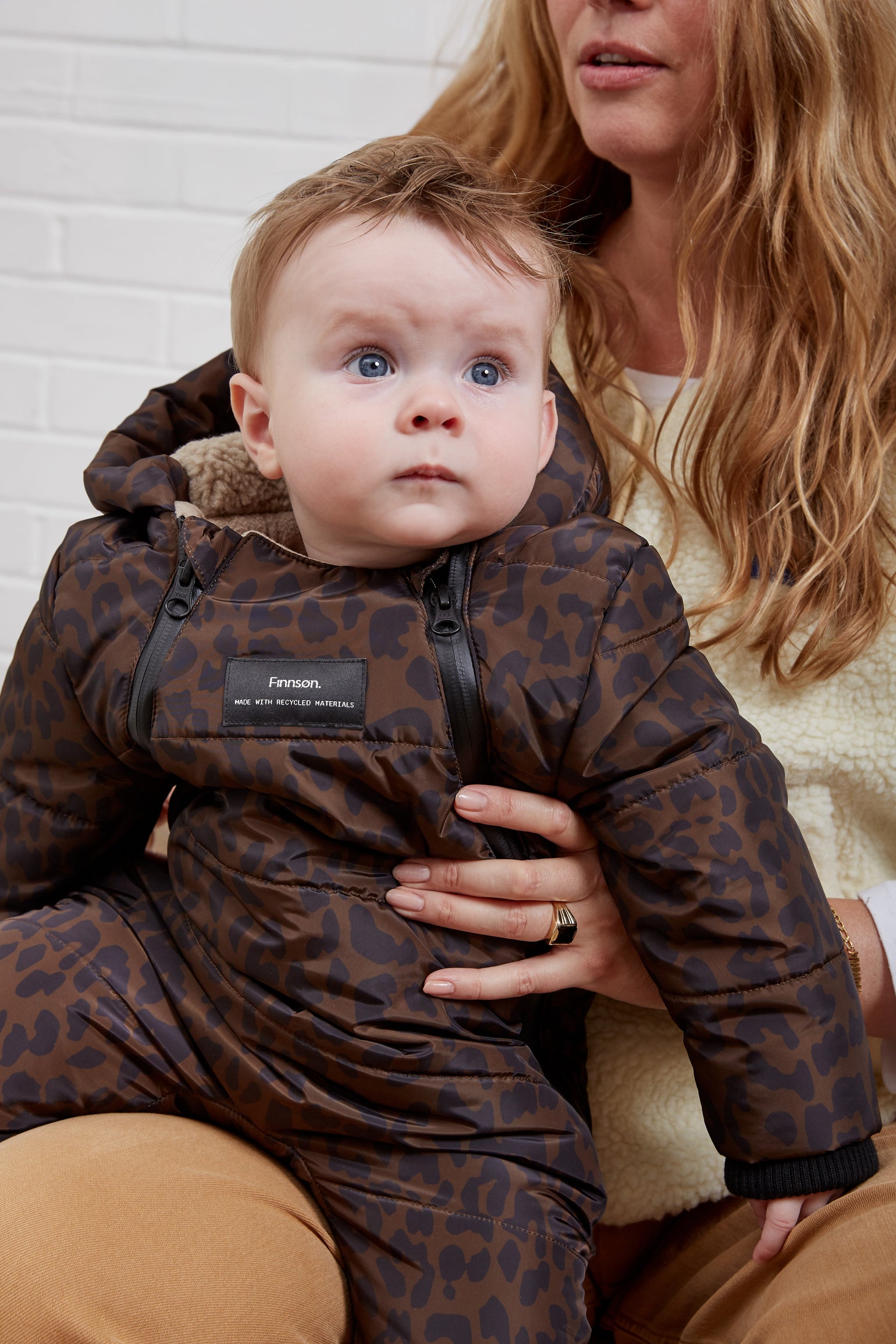 A baby wearing anders choco leopard print snow suit, held by a woman against a white brick wall background.